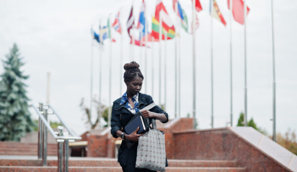 African student female posed with backpack and school items on yard of university, against flags of different countries.